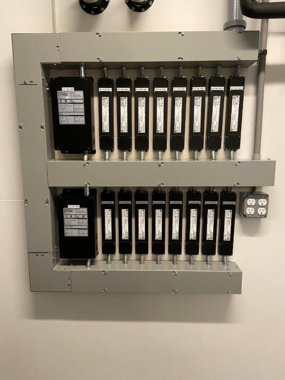 Gray electrical panel with black rectangular components and multiple fuses. White wall.