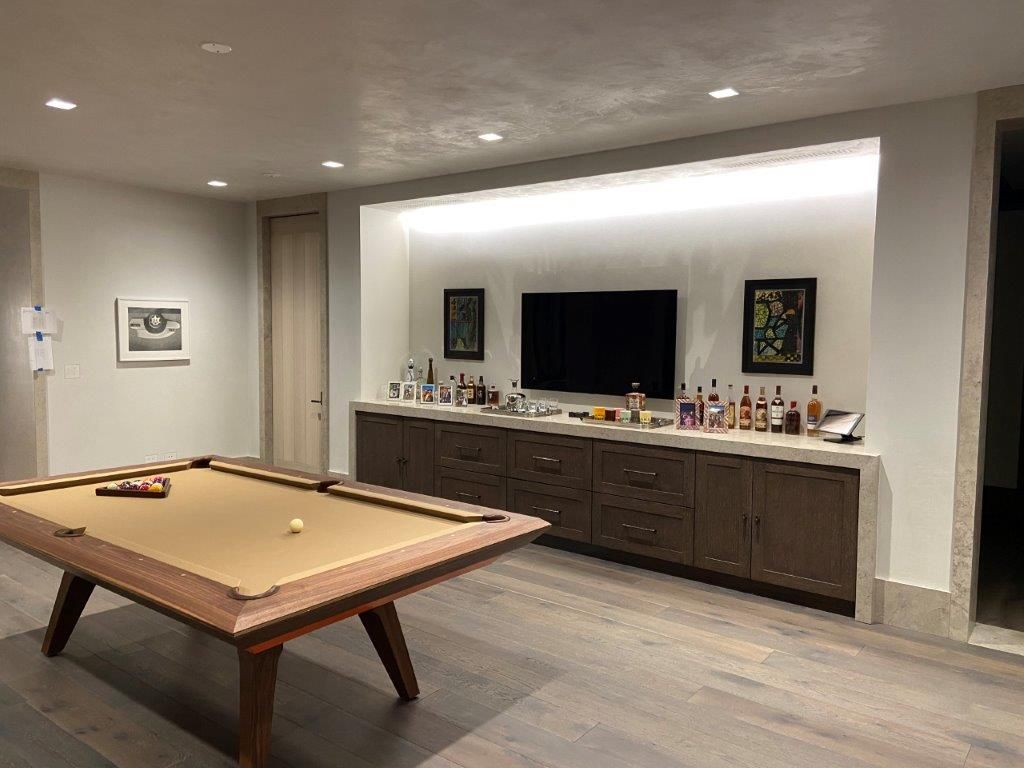 A modern game room with a pool table, built-in bar, and TV; neutral color palette.