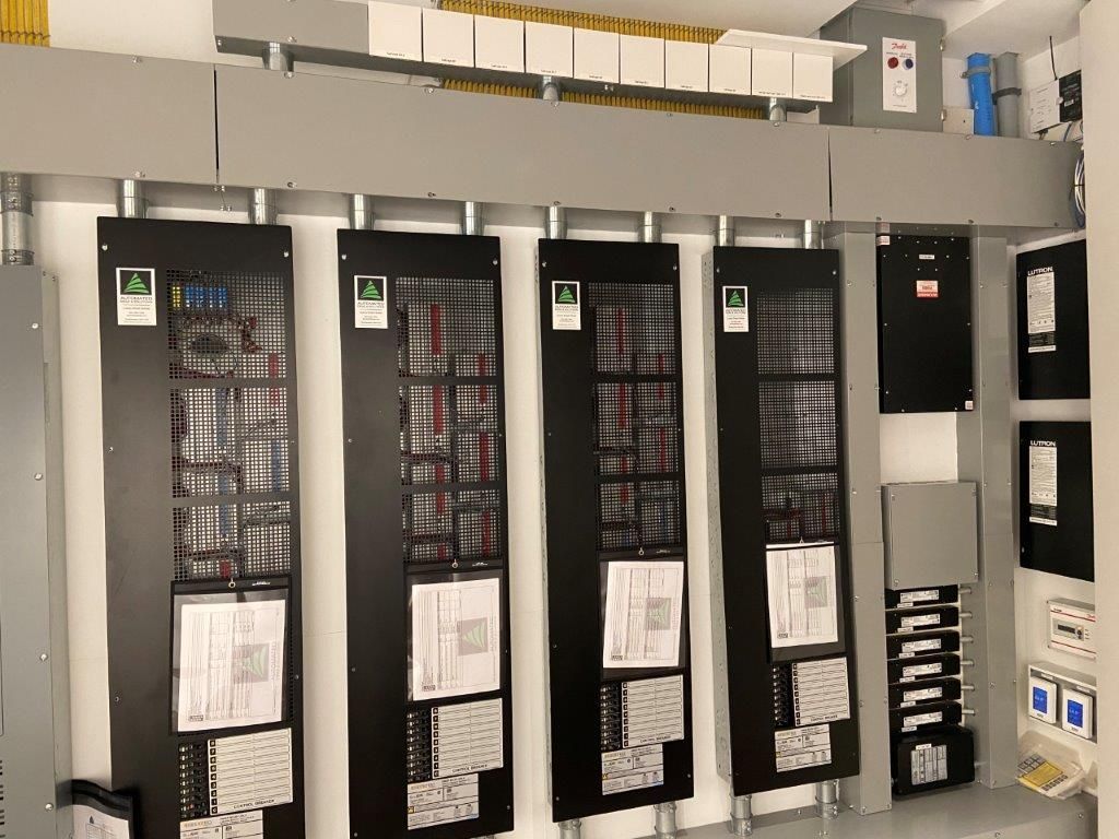 Electrical panels mounted on a wall. Several black panels with documentation and wiring. Grey enclosure above.