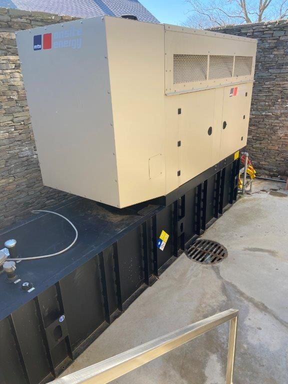 Large beige generator on a black fuel tank, set against a stone wall and concrete pad.