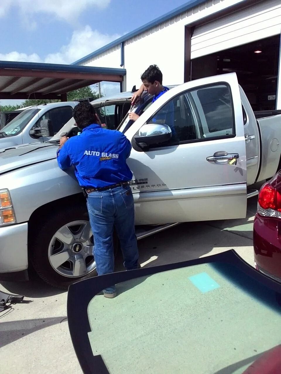 Two people installing a windshield on a silver pickup truck outside a repair shop.