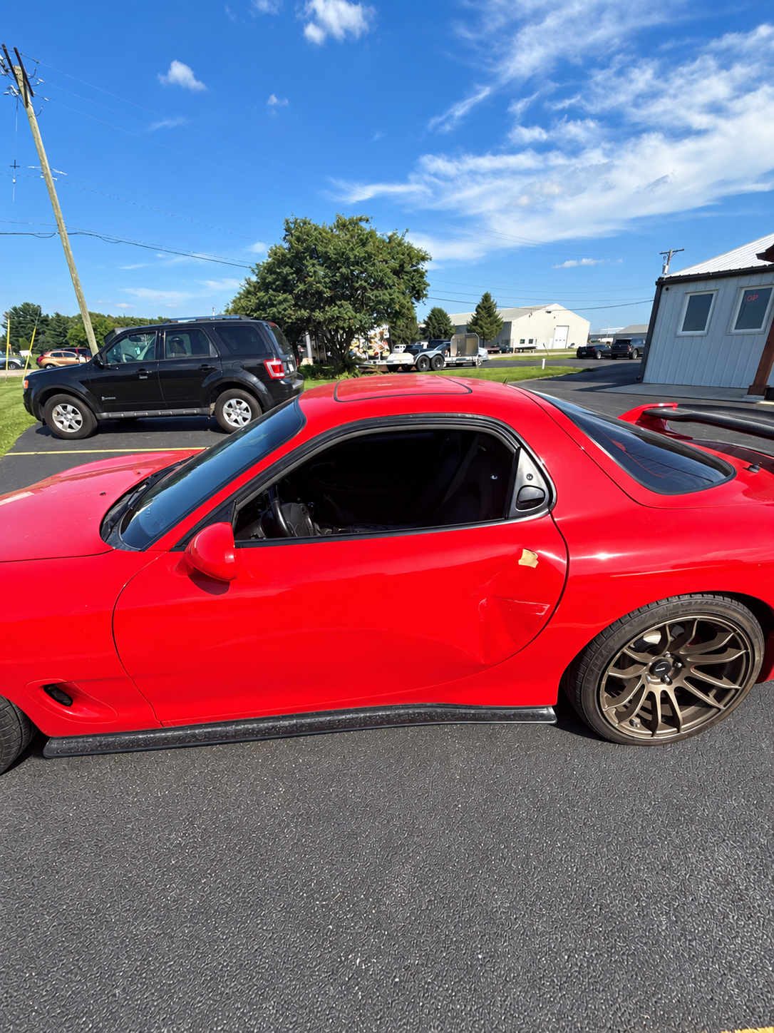 Red sports car parked on asphalt under a blue sky, with a black SUV and houses in the background