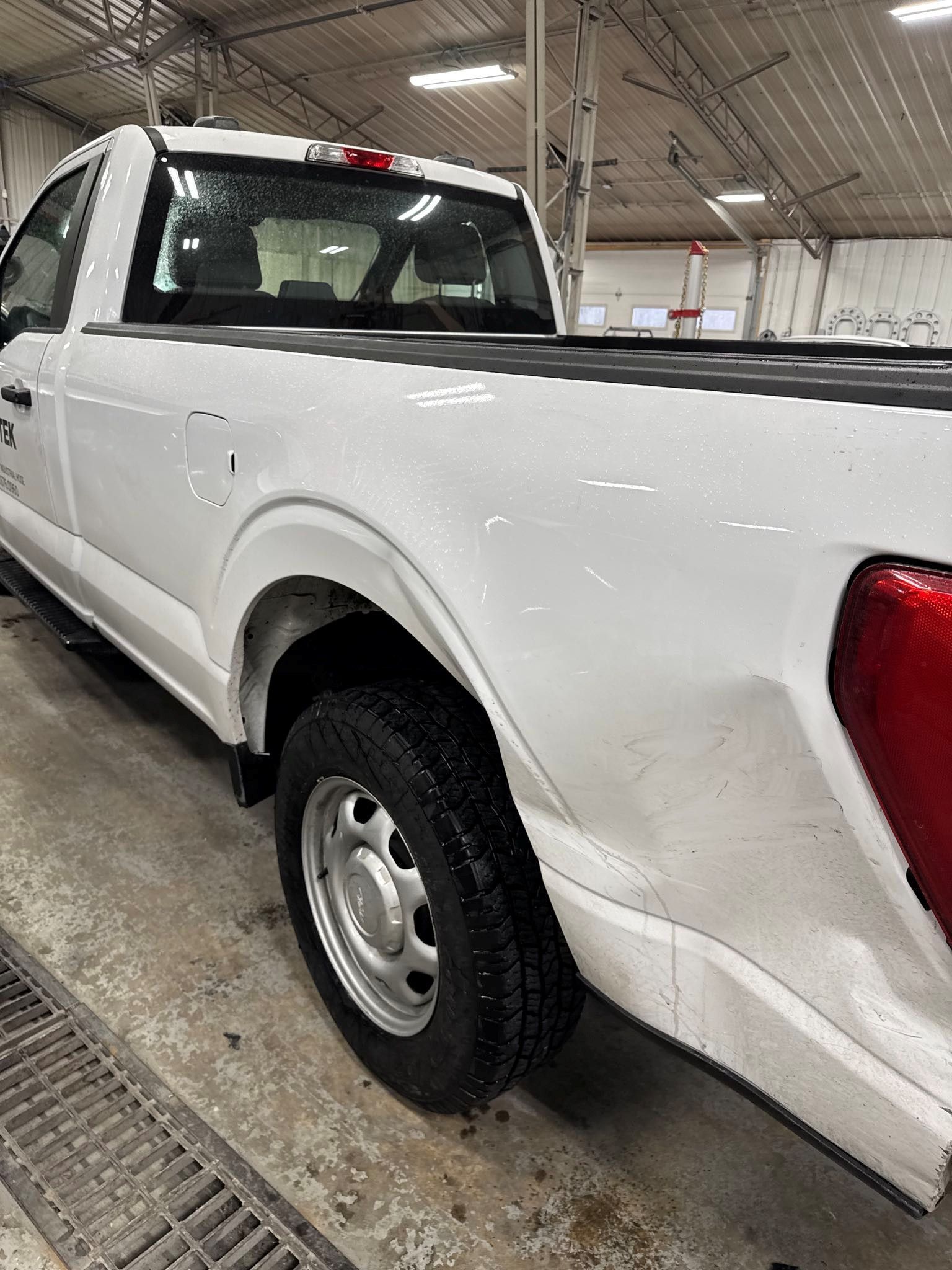 White pickup truck, rear view, in a shop, black tire, silver wheel, black bed cover, and red tail light - before
