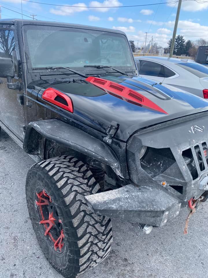 Black Jeep with red accents on the hood and wheels, parked outdoors on a cloudy day.