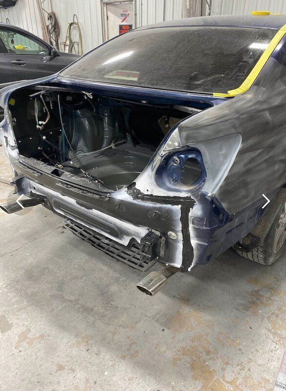 Rear of a blue car, undergoing bodywork in a garage; bumper removed, primed areas, tail light visible, exhaust pipes.