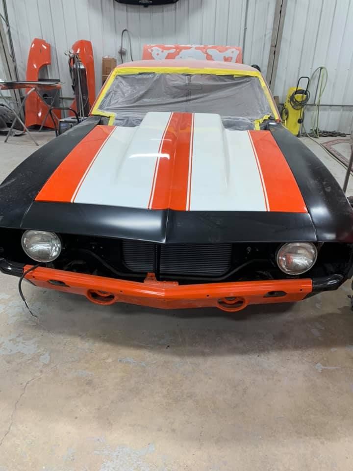 Black and orange classic car being worked on in a garage; hood with white and orange stripes.