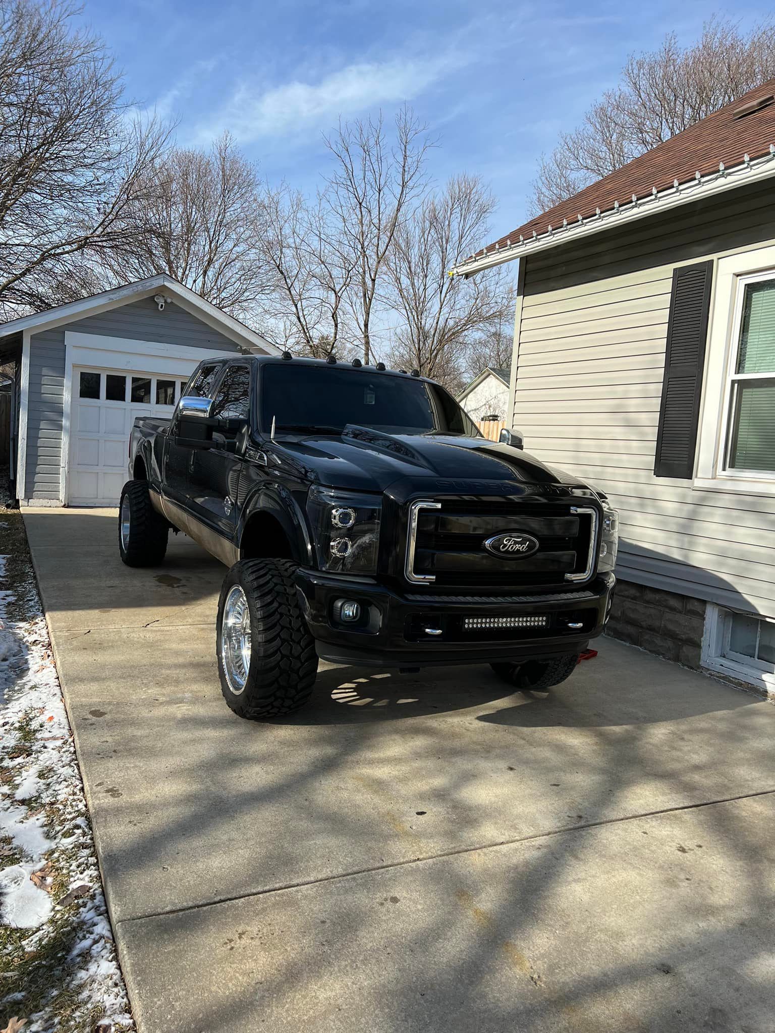 Black lifted pickup truck parked on a driveway in front of a gray garage and house on a sunny day.