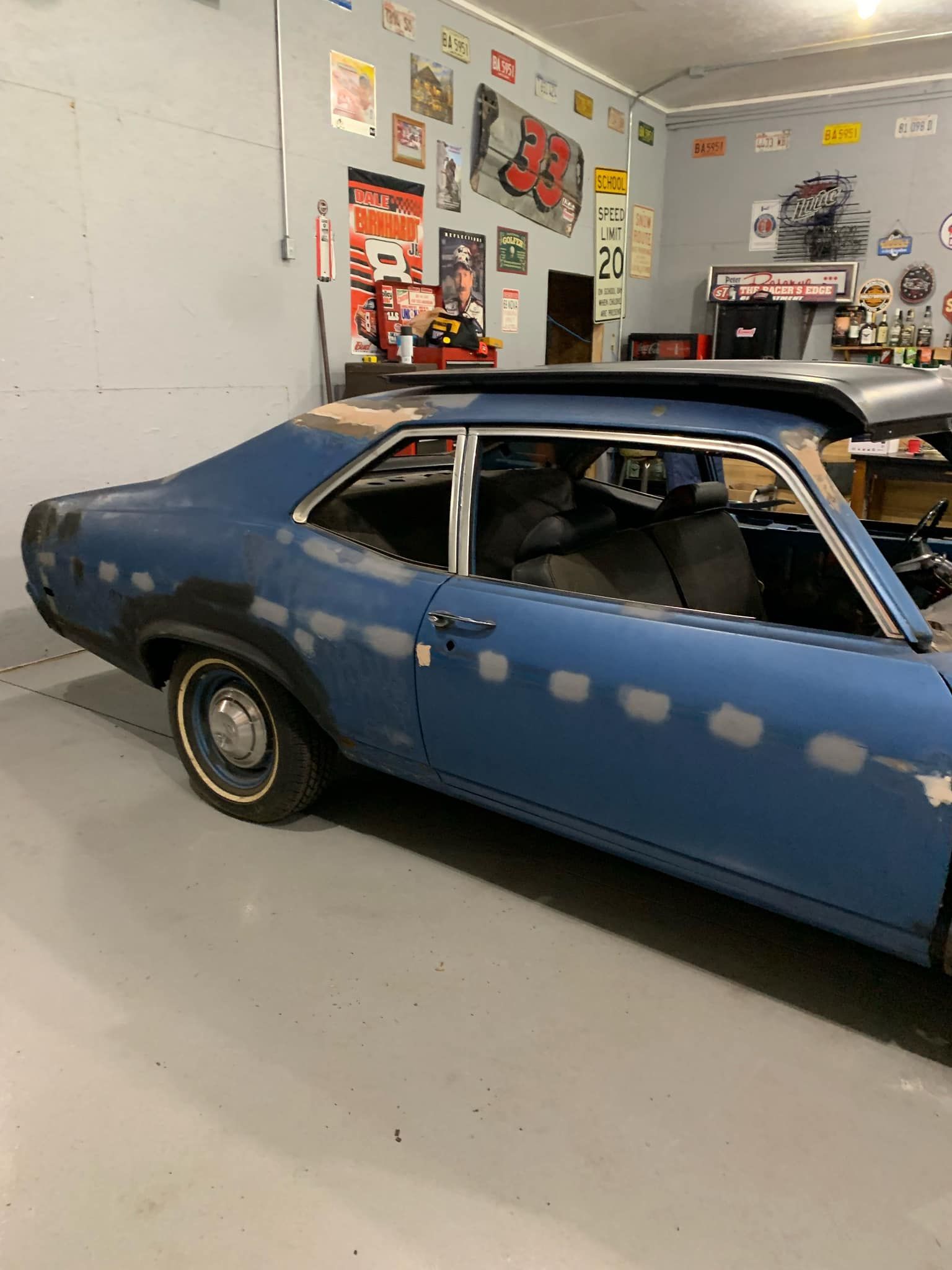 Blue classic car in a garage, undergoing restoration. Grey walls, signs, and tools are visible.
