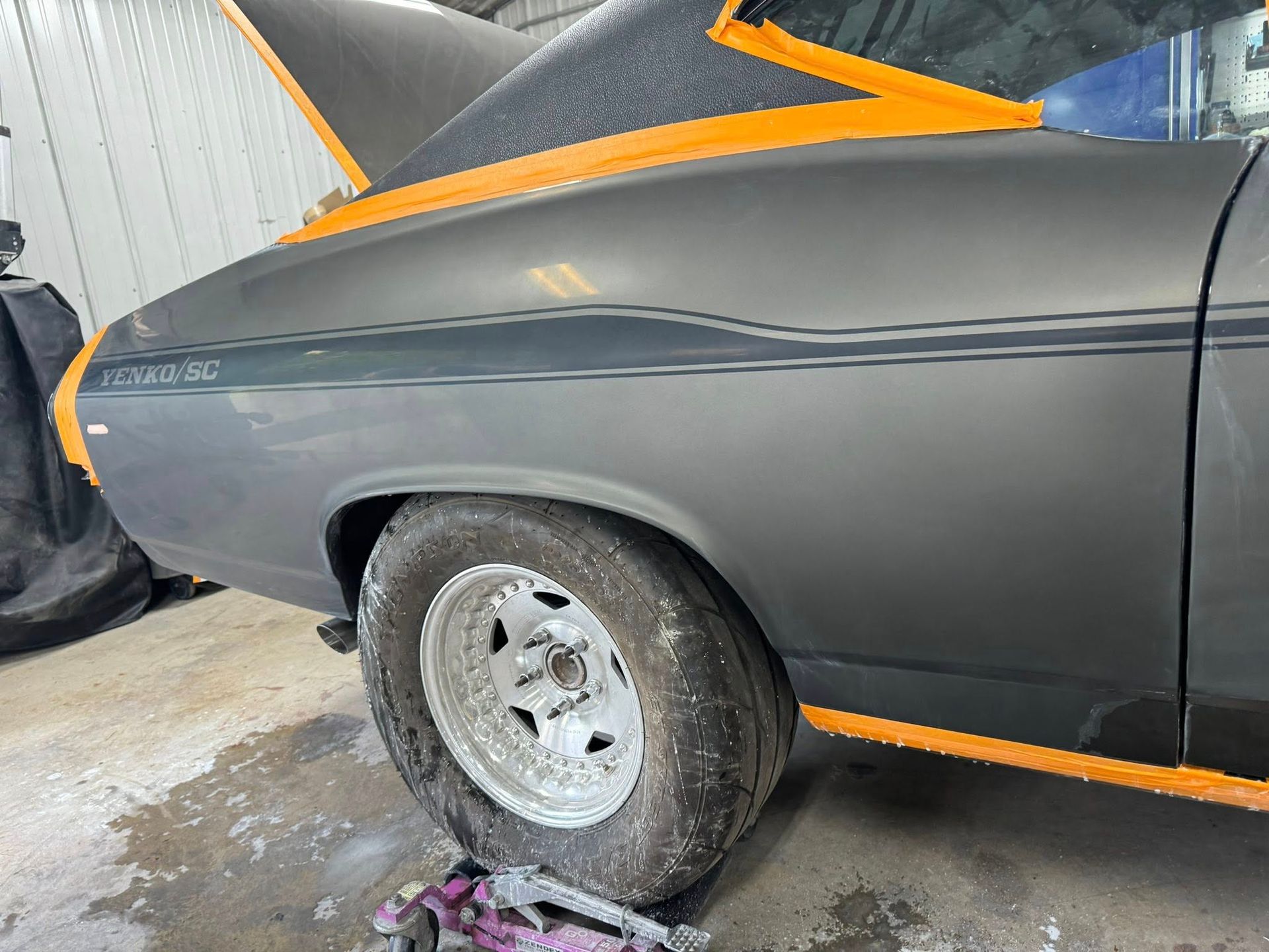 Gray classic car's rear quarter panel being prepped for paint, with orange tape and a racing stripe.