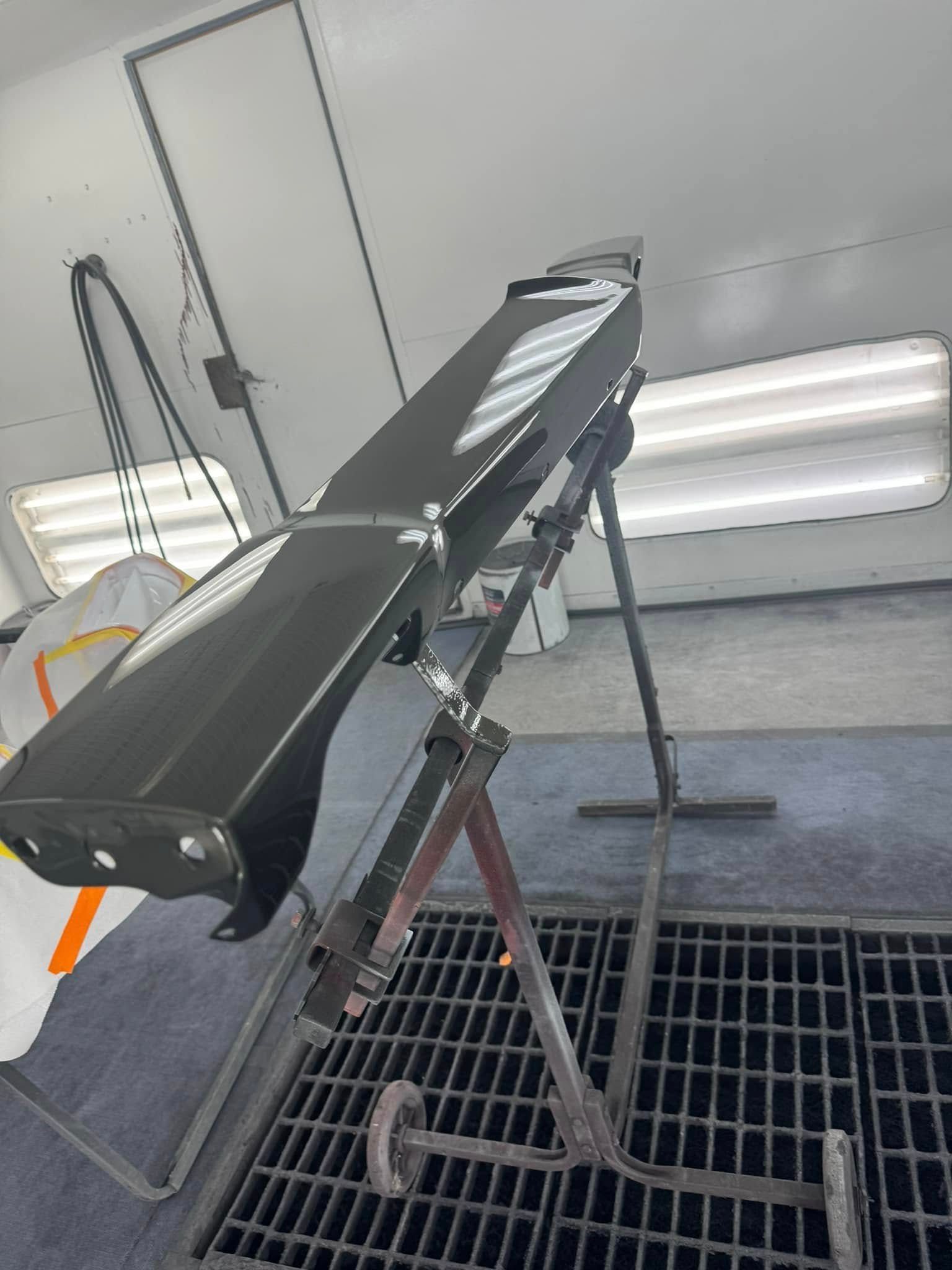 Black car part on a paint stand inside a paint booth with white walls and overhead lights.