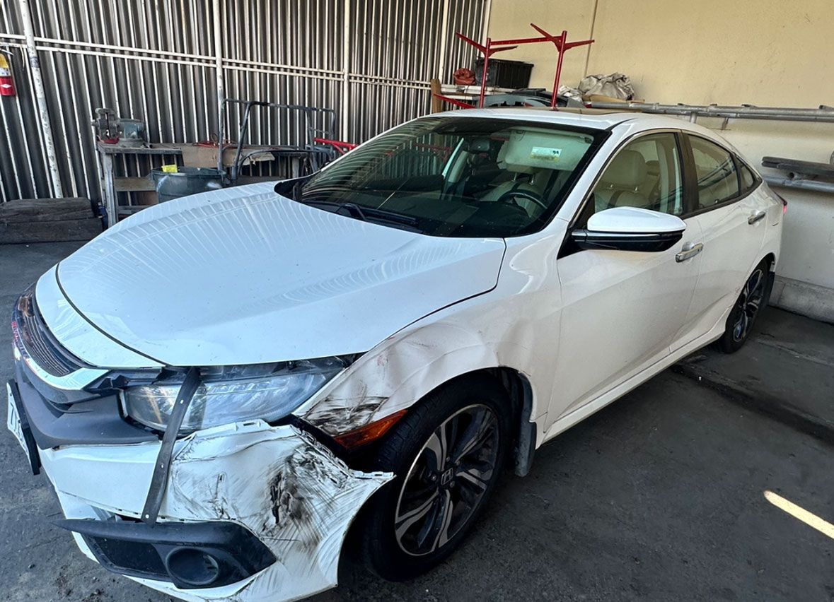 A white car with a damaged front end is parked in front of a building.