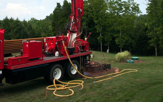 Red drilling rig boring a well in a grassy yard, trees in background.