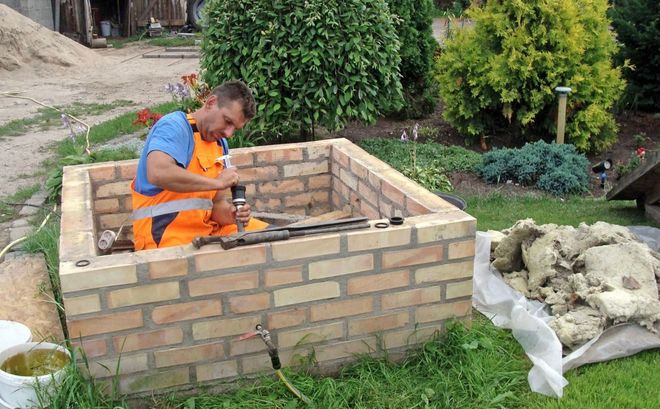 Man in orange overalls working inside a brick-lined square pit outdoors; tools nearby, garden backdrop.