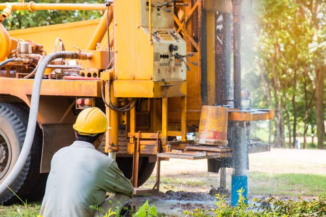 Yellow drilling rig extracting water, worker in yellow helmet observing, outdoors.