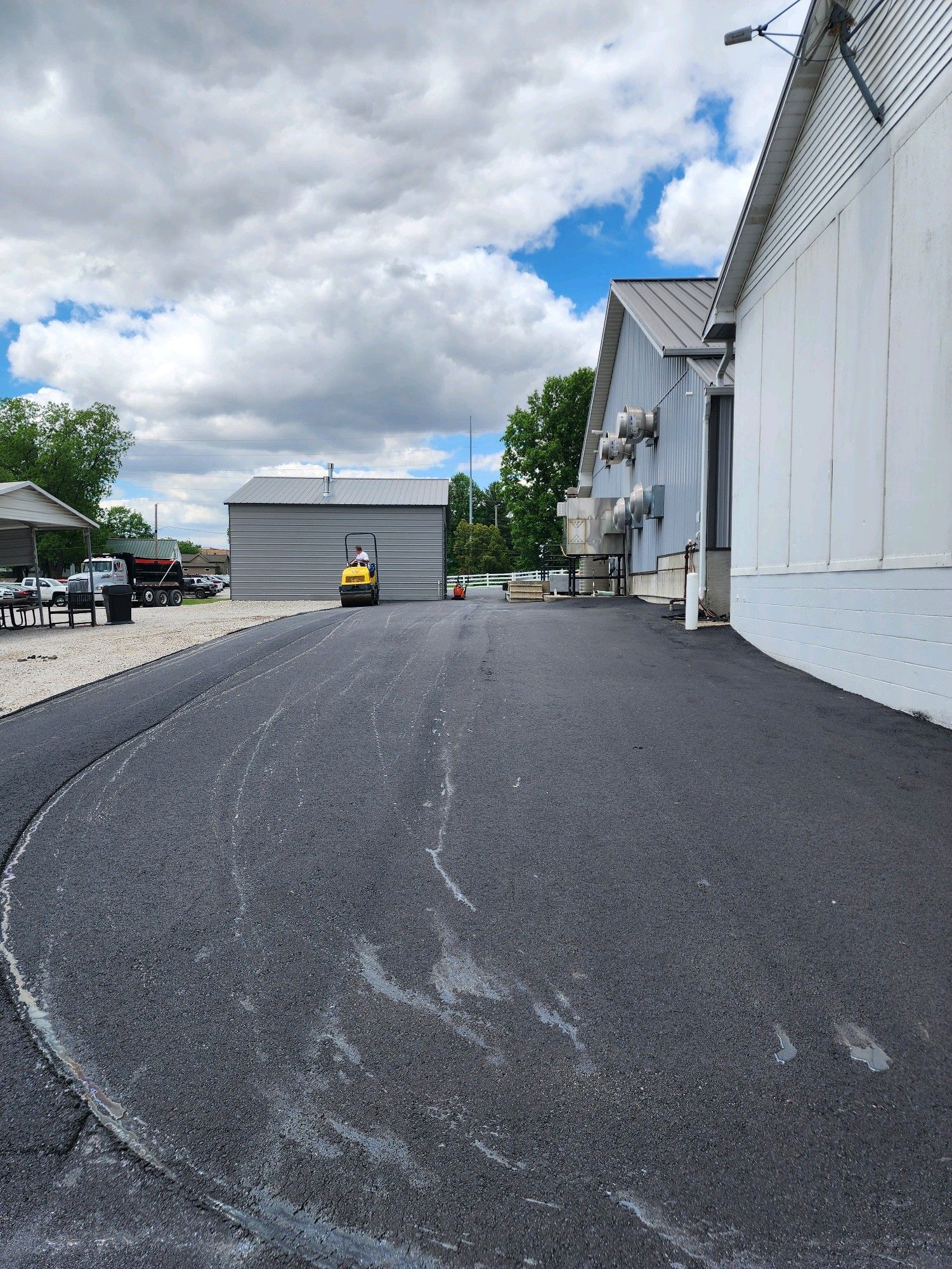 A yellow road roller sits on a freshly paved black asphalt surface between a metal building and a white structure.