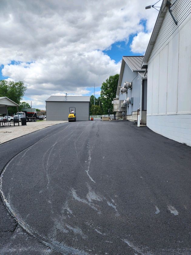 A yellow road roller sits on a freshly paved black asphalt surface between a metal building and a white structure.