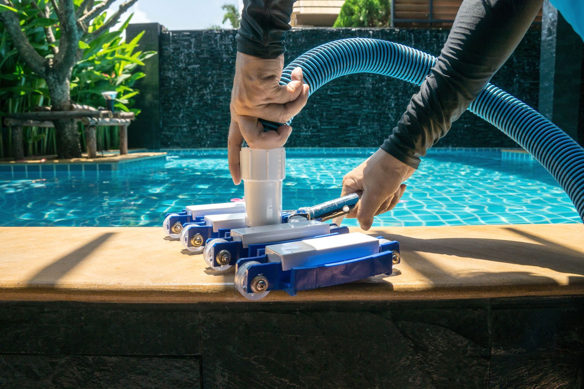 Person vacuums a blue-tiled swimming pool with a blue and white pool cleaner and hose, near a waterfall.