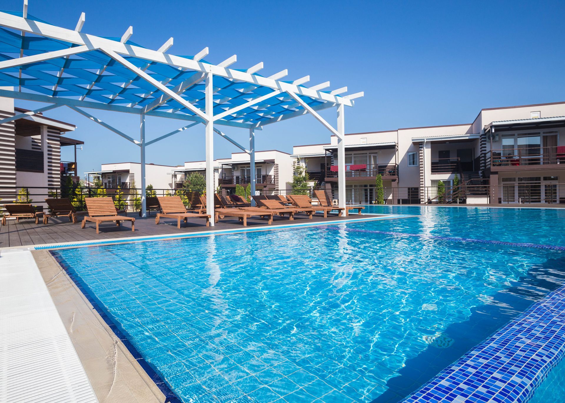 Pool with blue water and wooden lounge chairs under a blue pergola, with white buildings in background.