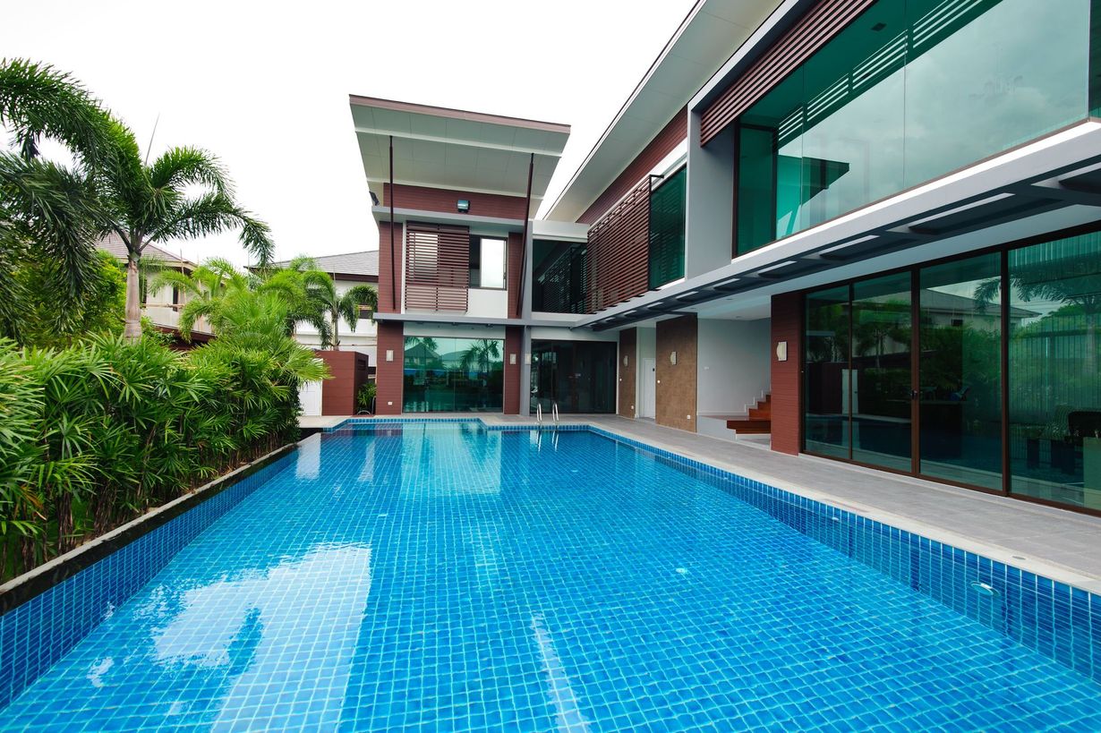 Modern house with a long, blue swimming pool in front. Large glass windows and green trees.