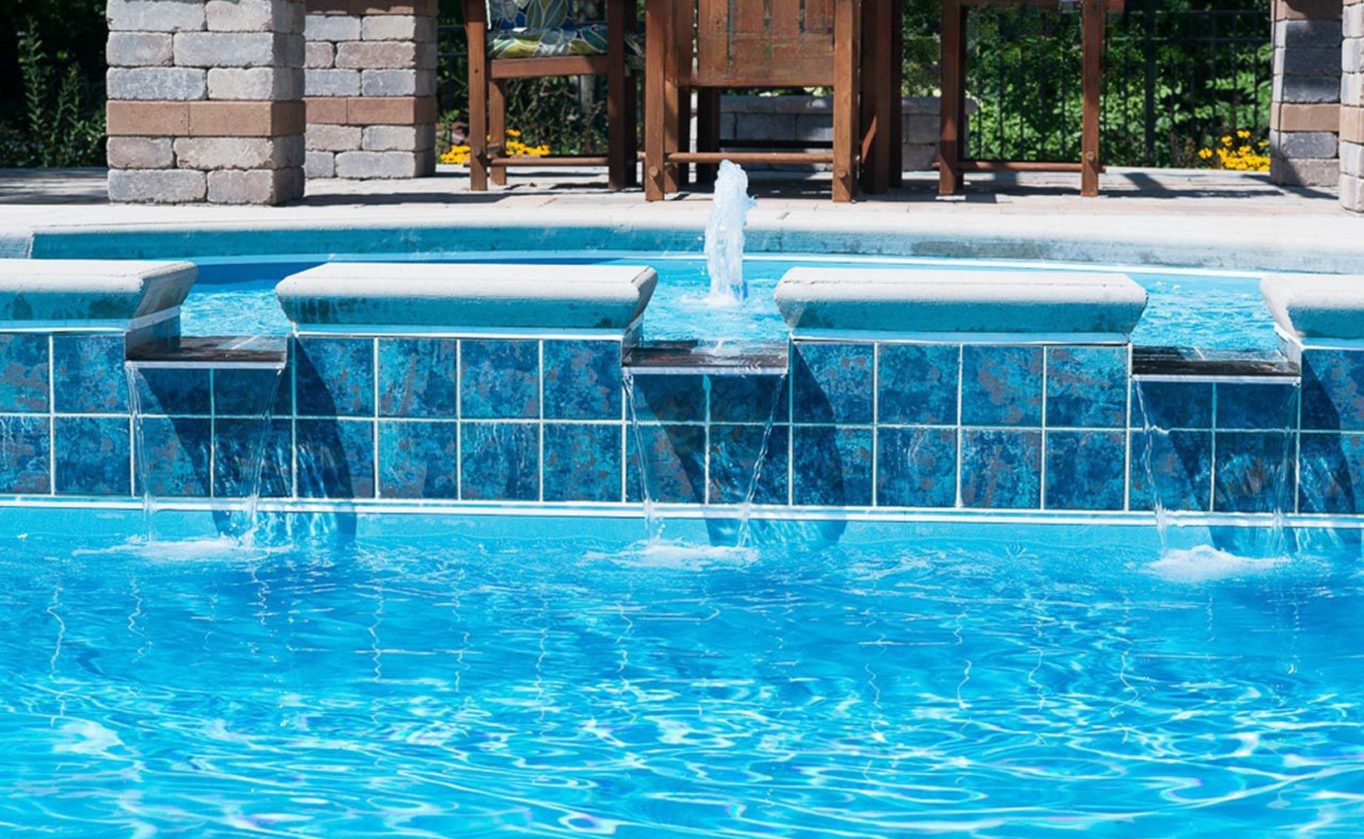 Blue swimming pool with cascading water over tiled ledge and lounge chairs in the background