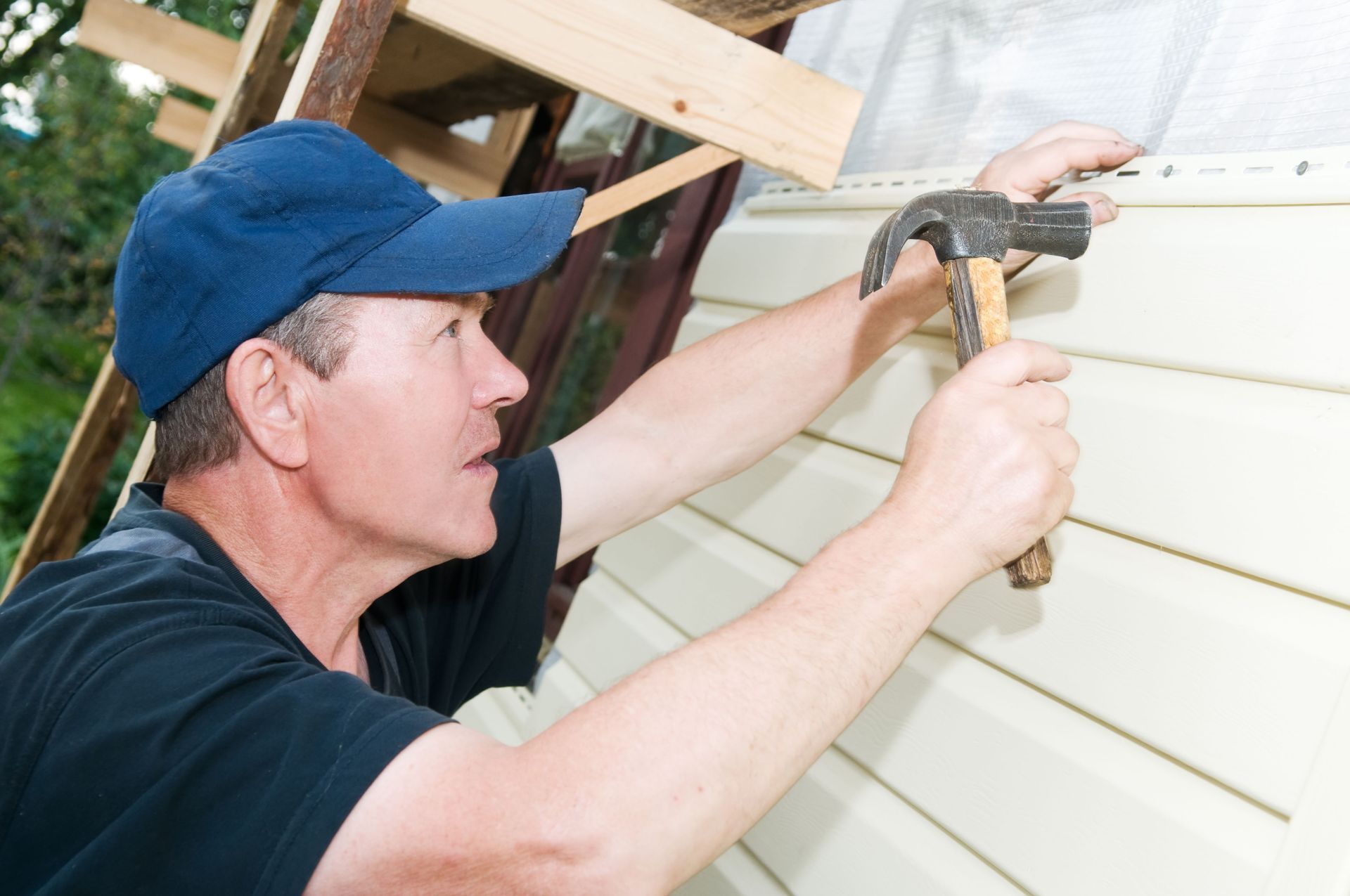 Man in blue cap hammers siding onto a building.