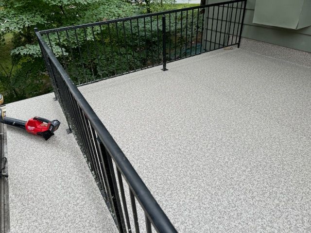 A light gray, speckled resin deck surface enclosed by a black metal railing, with a leaf blower resting on the floor.