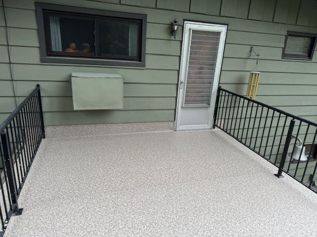 An outdoor deck with light-colored speckled flooring, black metal railings, a white door, and an external wall unit.