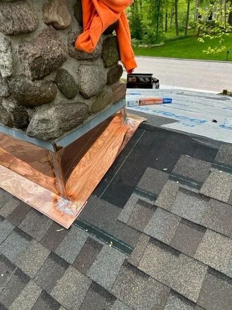 Copper flashing around stone chimney on a roof with shingle installation in progress. Orange cloth draped.