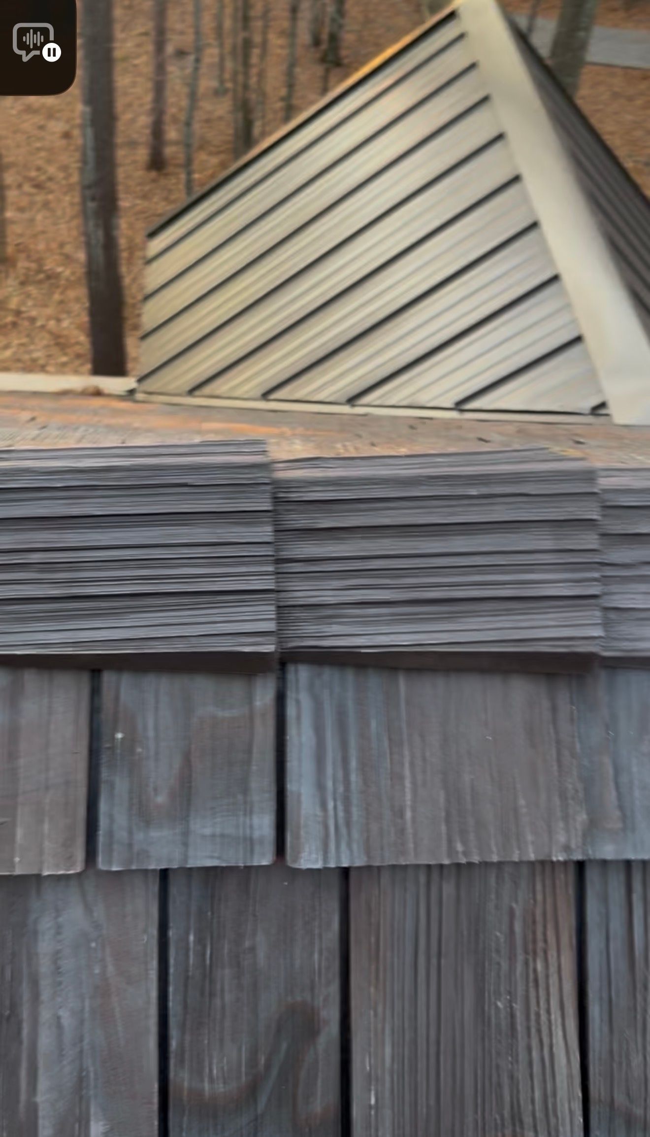 A close-up view of dark, textured wood shake roofing shingles in layers, with a blurred peaked roof in the background.