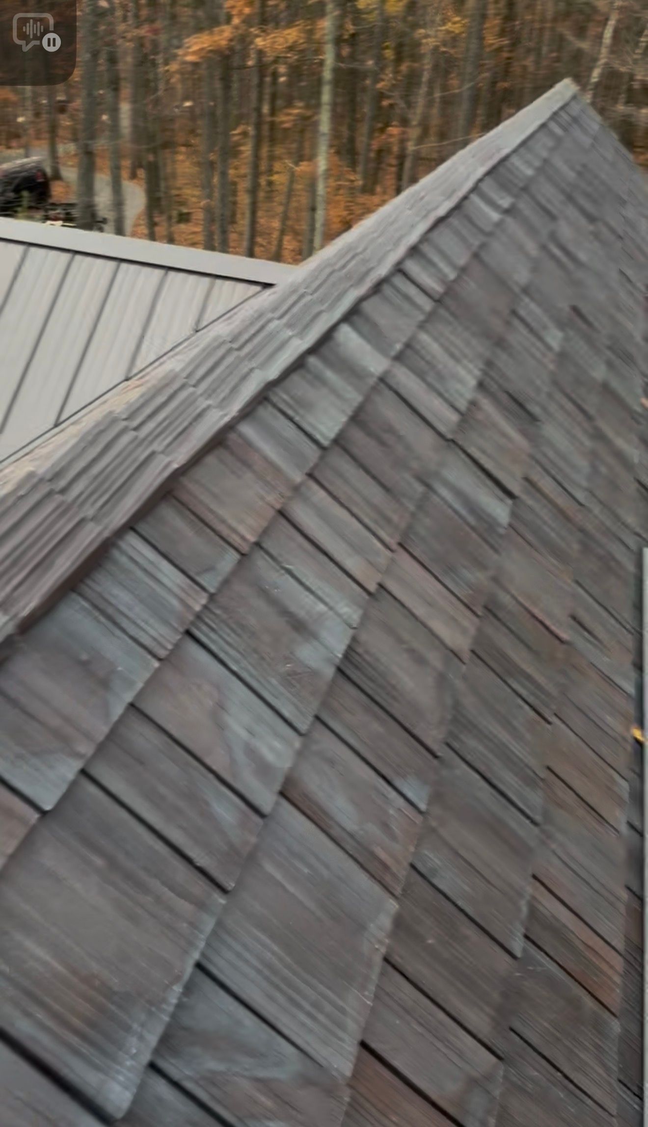 A steep roof with grey shingles meeting a metal-seamed section under a backdrop of autumn trees.