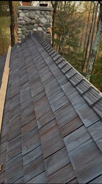 A high-angle view of a grey shingled roof with a stone chimney in the background and a line of trees nearby.
