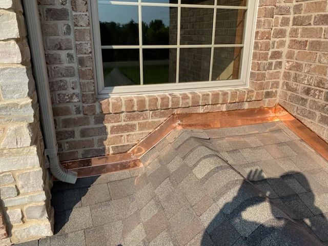 Newly installed copper flashing where a shingled roof meets a brick wall and window, with a downspout on the left.