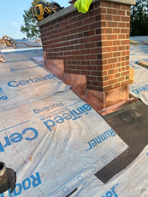 A brick chimney on a roof under construction, featuring newly installed copper flashing over the underlayment.