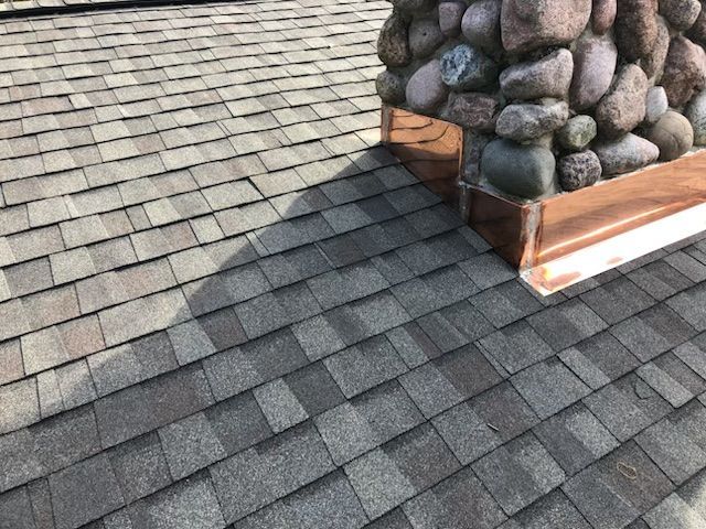 A stone chimney on a roof with asphalt shingles and copper flashing installed at the base.
