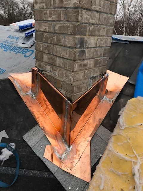 A copper chimney flashing installed on a roof with a brick chimney stack, showing soldered joints and base plates.
