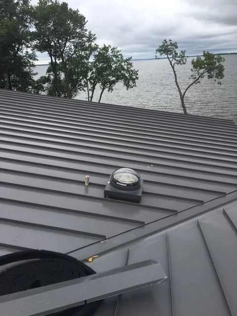 Gray metal roof with a skylight, trees, and water in the background under an overcast sky.