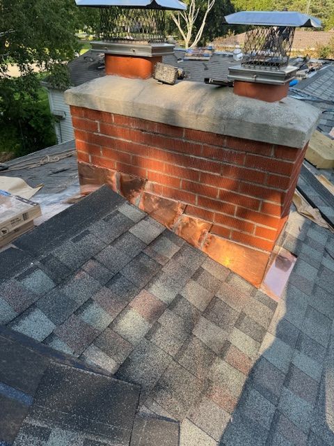A brick chimney with two metal caps sits on a sloped roof covered in multi-toned shingles during a roofing project.