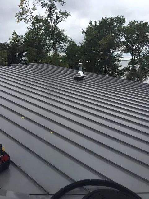 Grey metal roof with vent against a backdrop of trees.