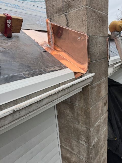 A copper flashing transition piece installed on a residential roof against a concrete block chimney.