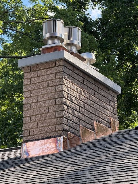 A brick chimney on a shingled roof with three stainless steel chimney caps against a background of green trees.