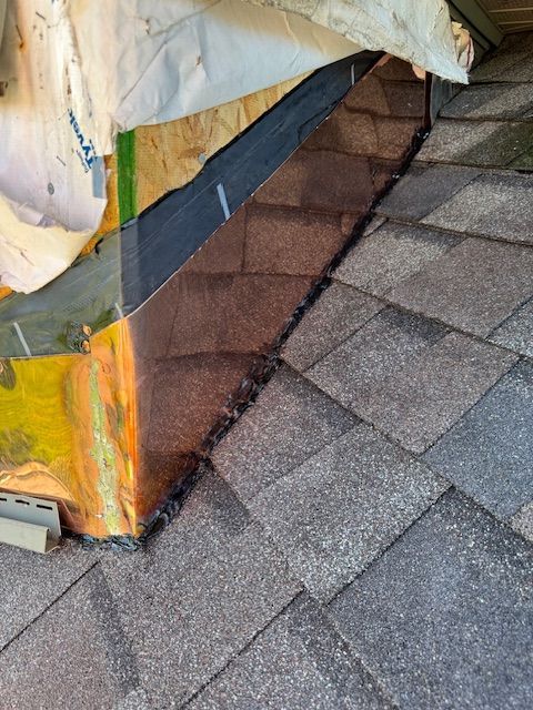 Close-up of residential roof shingles transitioning to a wall with brown flashing, black sealant, and construction wrap.