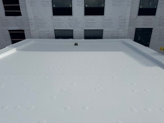 A flat, white roof surface under construction, viewed from above, with the exterior wall of a building in the background.