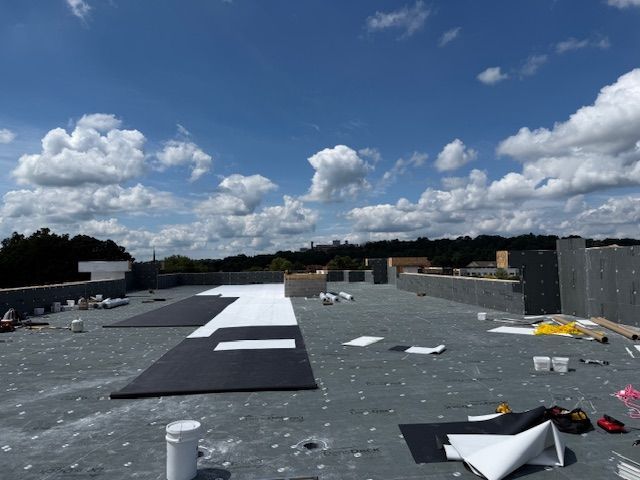 A rooftop under construction with dark and white insulating panels laid out on a gray surface under a blue cloudy sky.