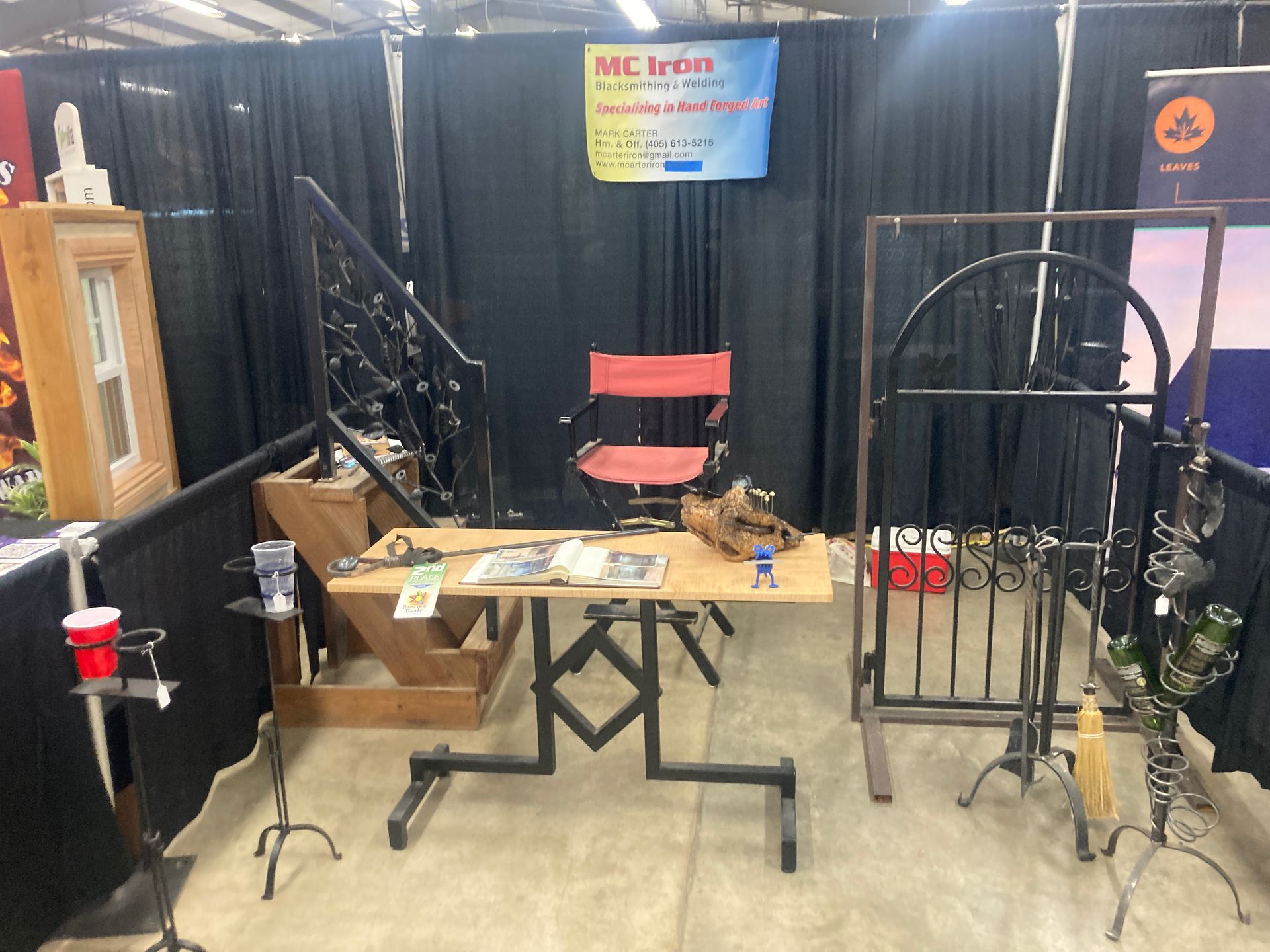 A trade show booth featuring iron railings, a metal table with a wooden top, a red chair, and decorative iron displays.