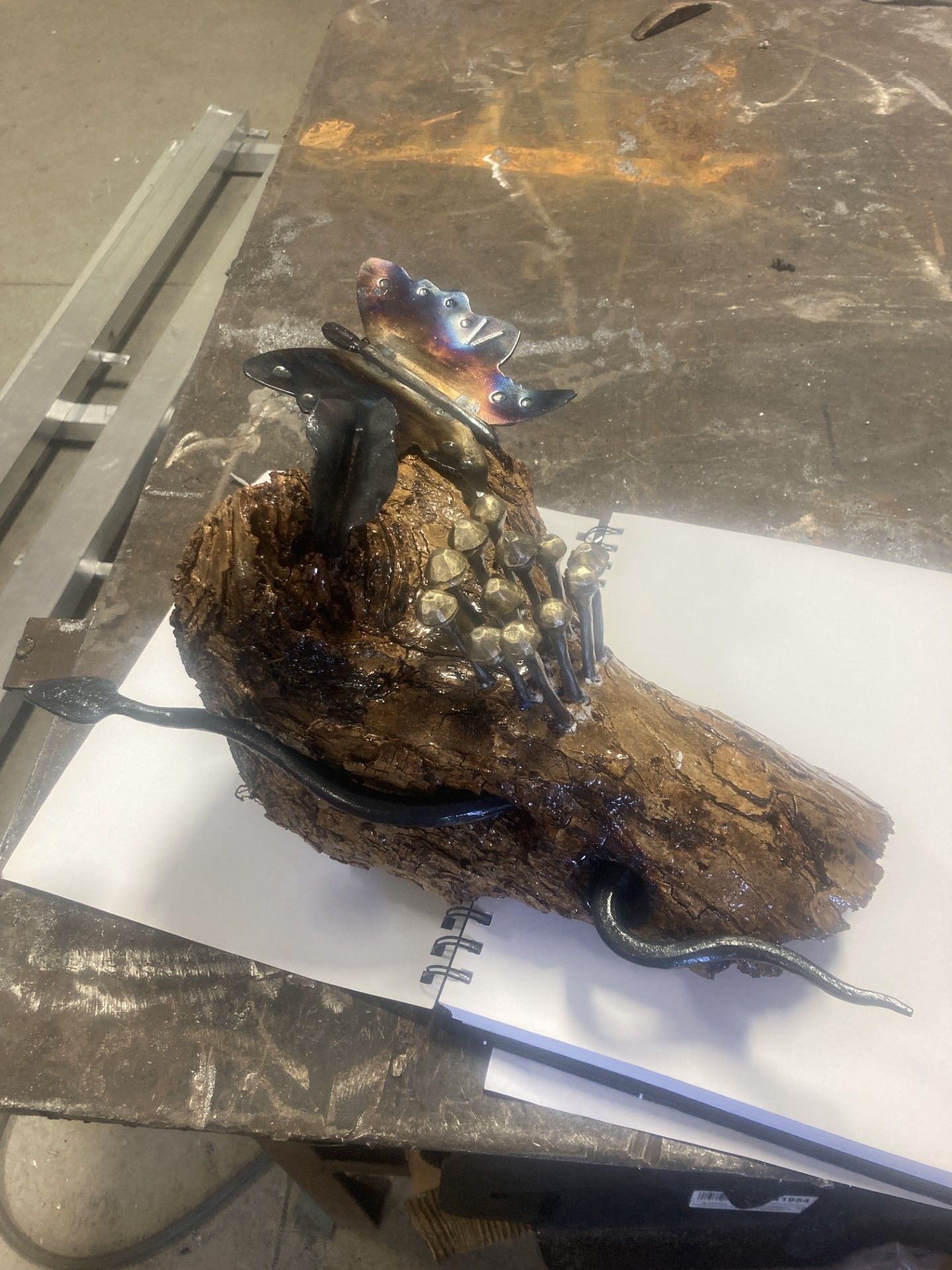 A metal-welded sculpture featuring a snake crawling over a textured, rock-like form with a metallic flower on top.