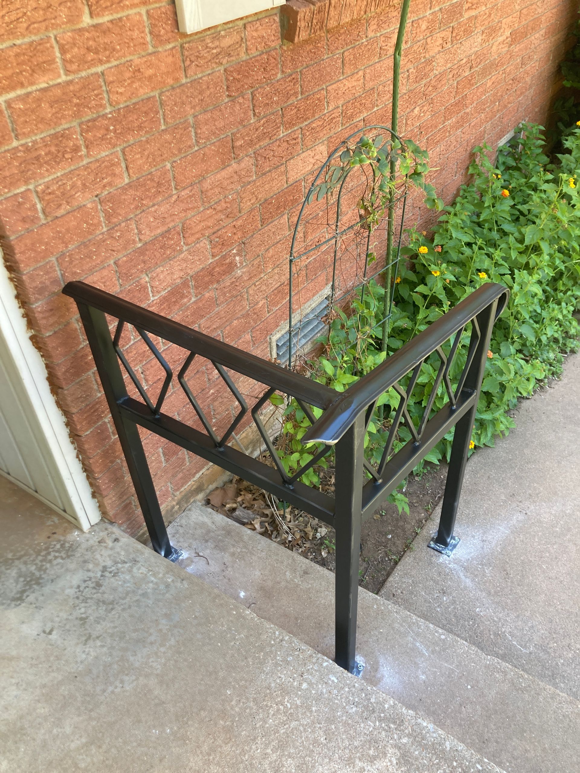 Black metal handrail and step barrier beside a brick wall, next to concrete steps and greenery.