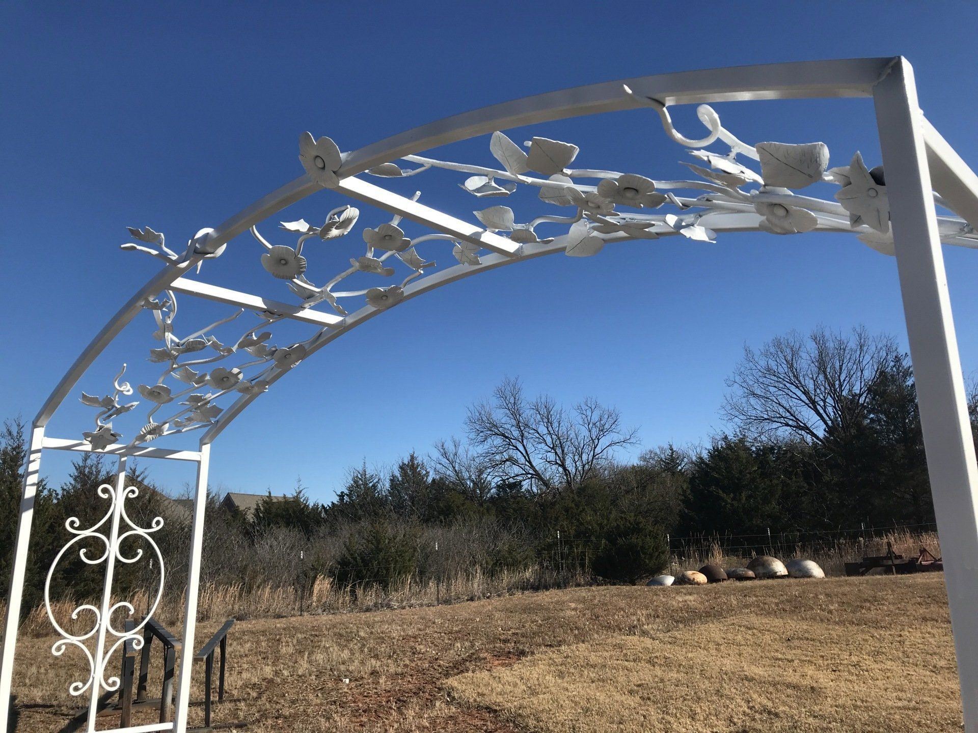 White metal garden arch with floral details against a blue sky, in a yard.