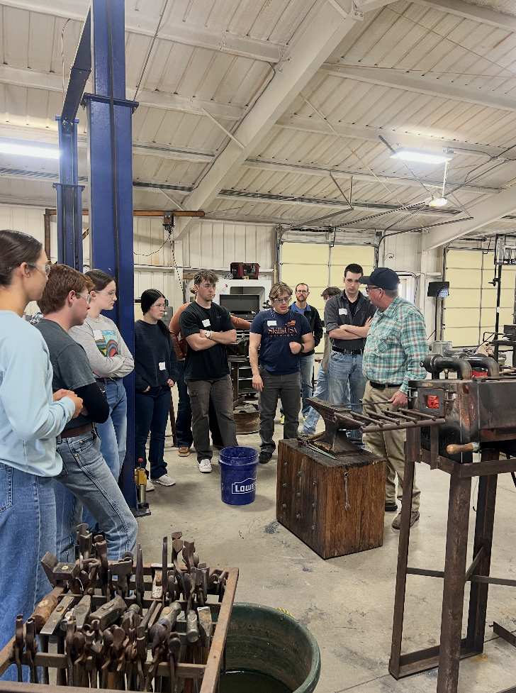 People gathered in a workshop, listening to a man near an anvil. Tools and equipment surround them.