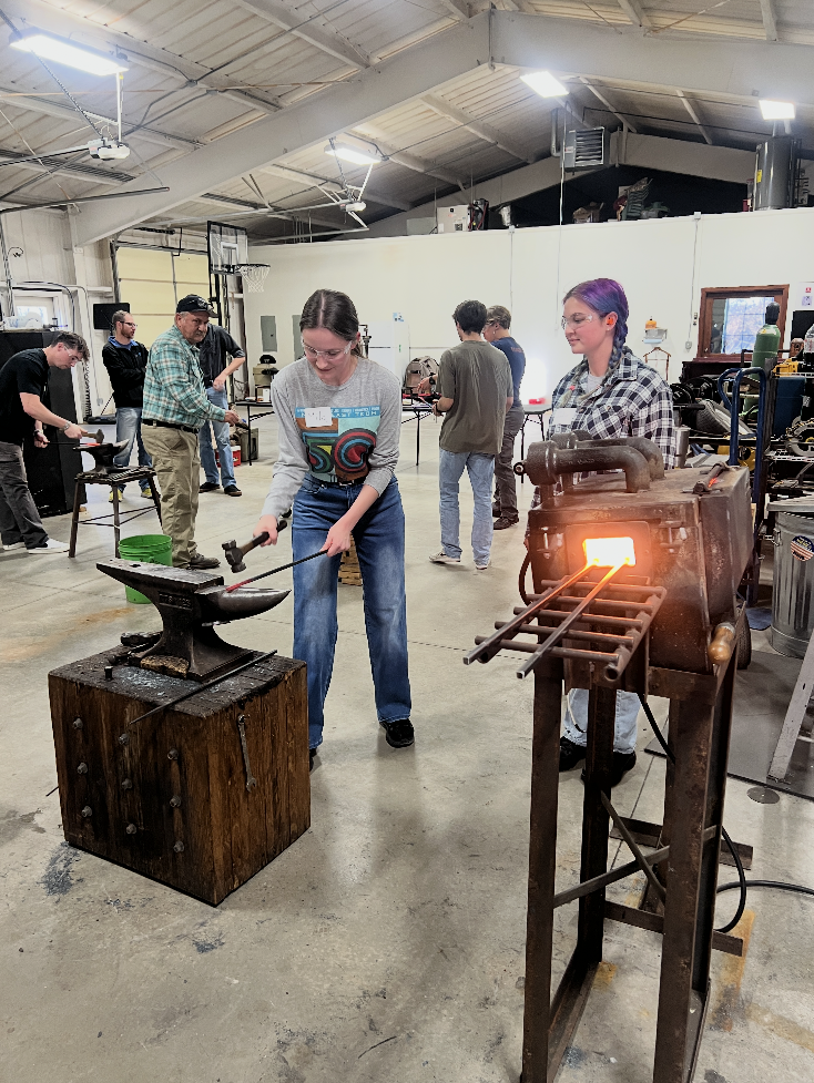 People at a blacksmithing workshop. One person hammers metal on an anvil; another attends a glowing forge.