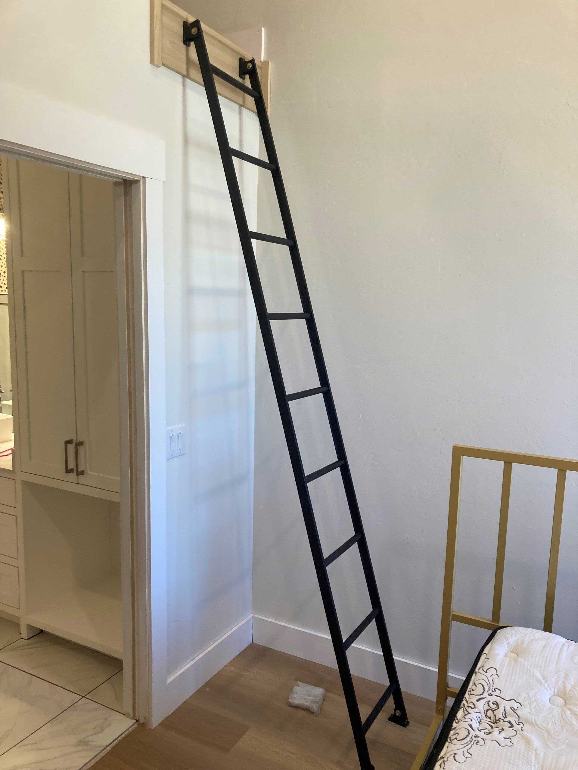 Black metal rolling ladder mounted on a white wall in a room. Adjacent to a white cabinet.